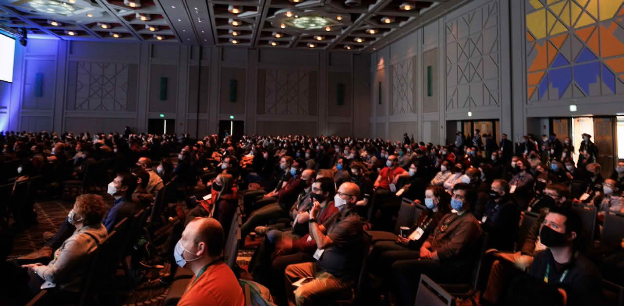 A large audience seated in a conference hall with a stage and screen in the background