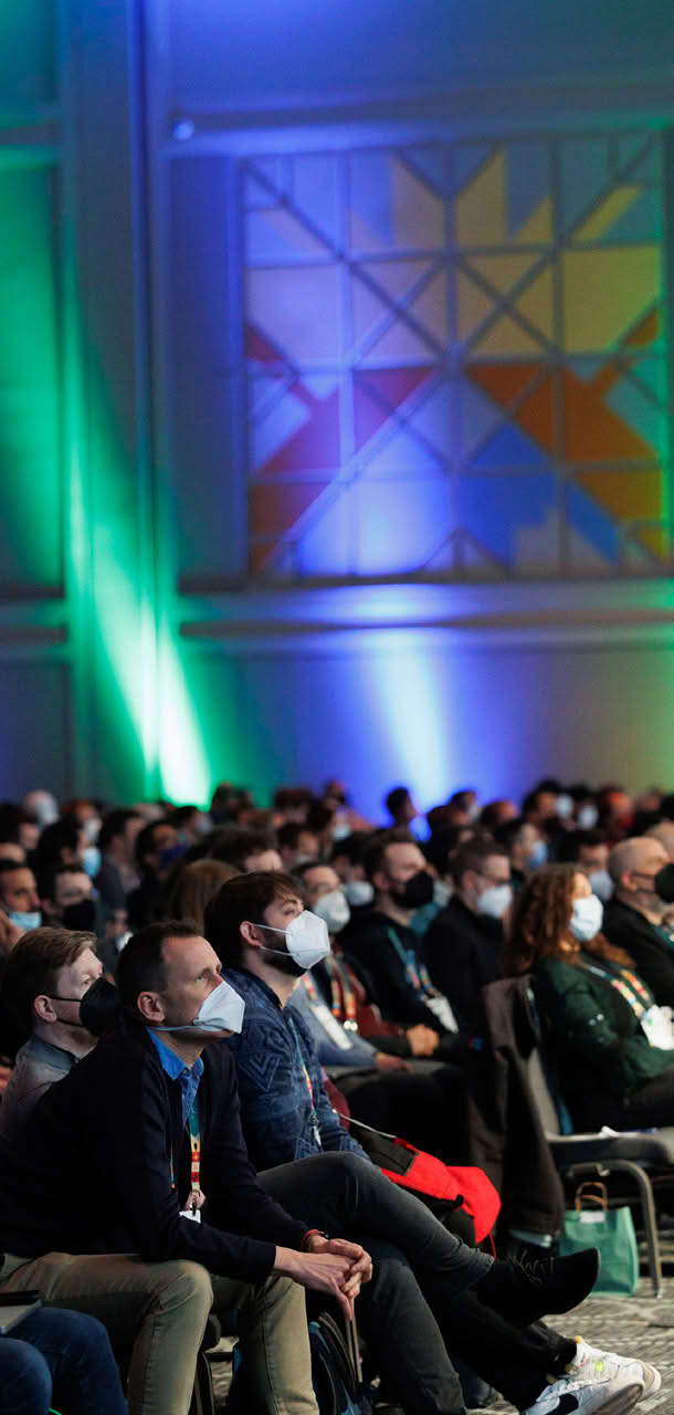 A large audience sitting in a conference hall, with colorful lights and geometric patterns on the walls